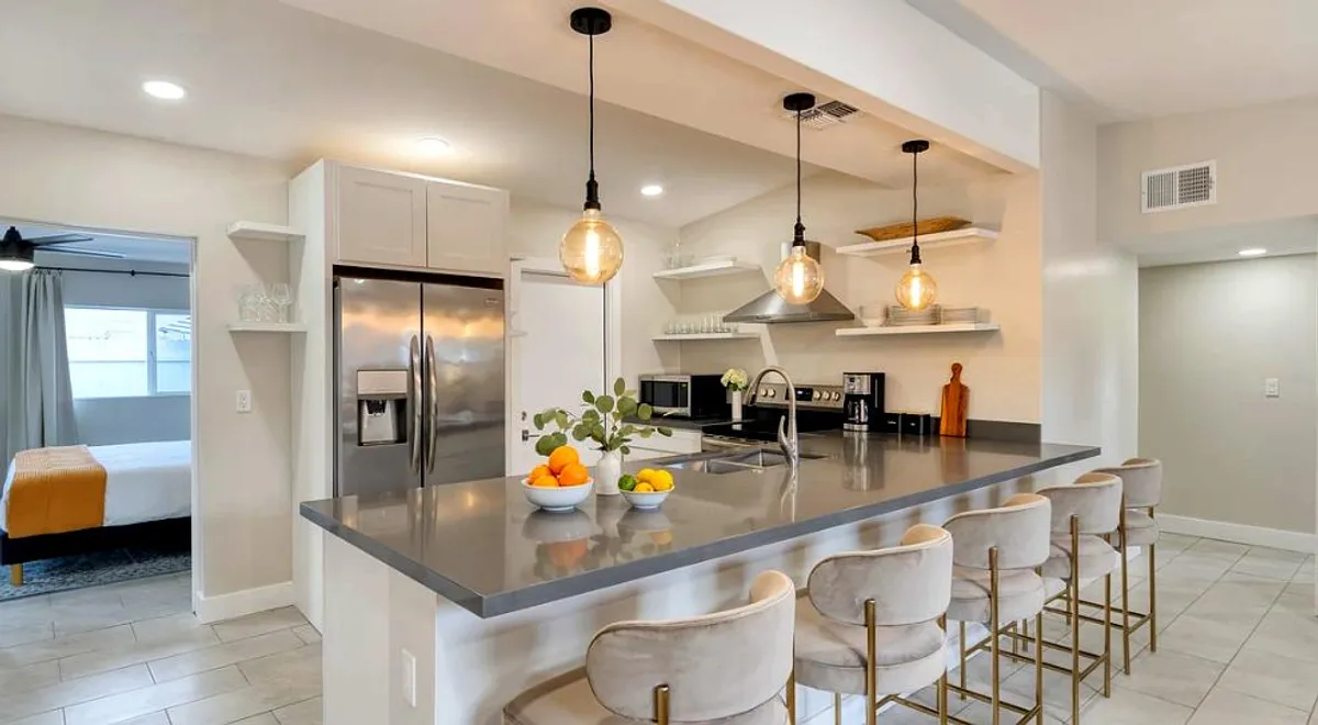 A contemporary kitchen is the star of this image, featuring a spacious island with pendant lights, stainless steel appliances, and a welcoming fruit bowl on the counter. The kitchen is designed for both functionality and style, encouraging guests to gather, cook, or enjoy casual meals.