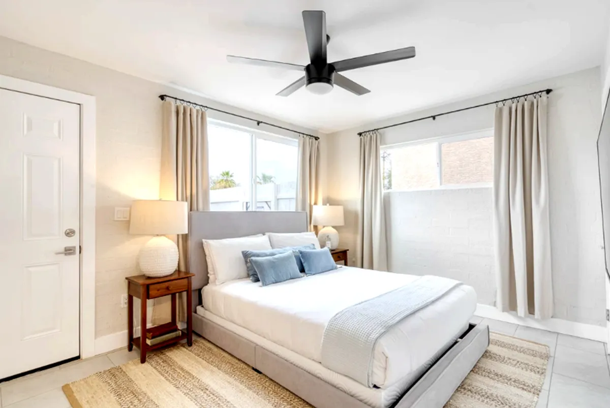 A serene bedroom featuring a neatly made bed with cozy blankets and pillows, table lamps on either side, a window allowing natural sunlight to stream in, and a ceiling fan above. The room exudes calm and comfort, ideal for rest and relaxation.