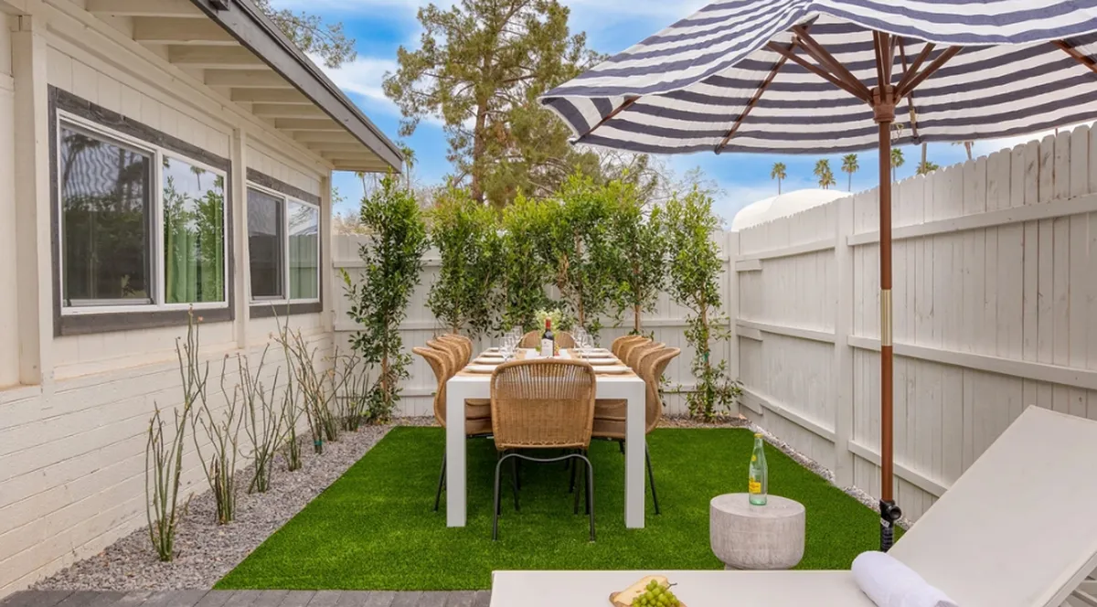 An inviting backyard scene featuring a dining table with an umbrella, surrounded by lush greenery and a comfortable lounge chair. The setup suggests both relaxation and social outdoor meals in a private, family-friendly space.