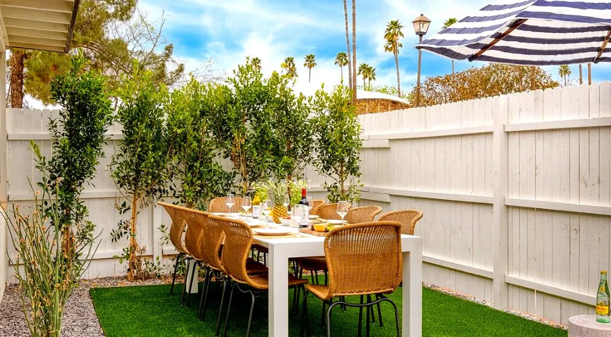 A cheerful outdoor dining area with a long table surrounded by woven chairs, lush greenery, and a bold striped umbrella. The setting is perfect for gathering, dining under the stars, or lingering over brunches in the Arizona sun.