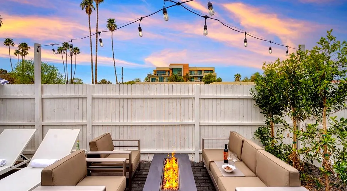 An exterior space with cozy lounge seating encircling a fire pit, enhanced by string lights, palm trees, and a landscaped view. The area is perfect for relaxing, socializing, or enjoying evening gatherings as the sun sets.