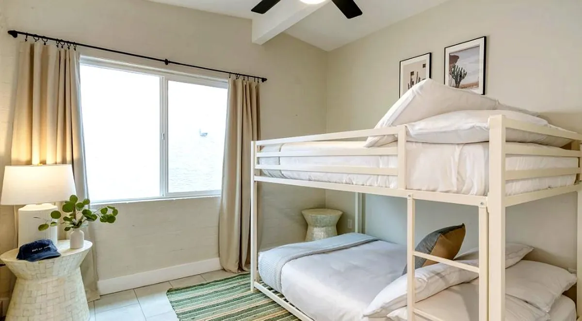 A cozy and playful bedroom designed for kids or friends, featuring a bunk bed with neatly arranged bedding, a window draped with curtains, a bedside lamp, and a decorative plant. The ambiance is inviting and perfect for creating lasting memories.