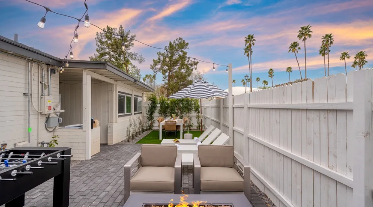 An inviting backyard scene featuring a cozy fire pit, outdoor seating, a table with umbrella, palm trees, and festive string lights. The ambiance is perfect for gatherings, post-game celebrations, or relaxing after a day out.