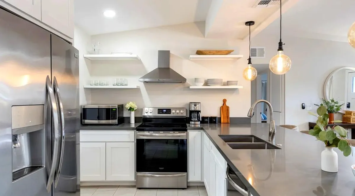 This image highlights another angle of the kitchen and dining area, showcasing the open shelving, stainless steel appliances, kitchen island, and pendant lighting. There's a clear view of spaces where people can gather for meals or conversation, reinforcing the interconnected design.