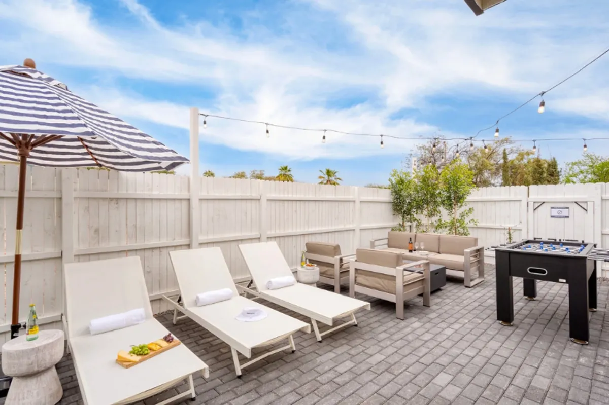 A spacious backyard with comfortable lounge chairs lined up beneath a large umbrella, a foosball table ready for play, and string lights overhead. The area blends spaces for sunbathing, playful games, and group gatherings, exuding a leisurely, inviting vibe.