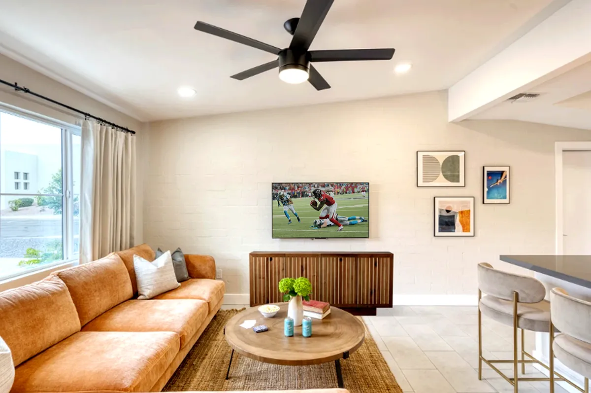 This image showcases an inviting living room with an orange sofa, a large flat-screen TV, a round coffee table, and decorative artwork. The space feels casual yet refined, inviting relaxation or social gatherings. Natural light filters in, highlighting the open layout and making it ideal for movie nights or enjoying a cup of coffee.